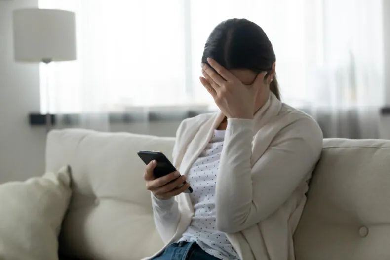 Woman sit on sofa holding smartphone cover face with hand feels scared and suffering from working with the insurance company